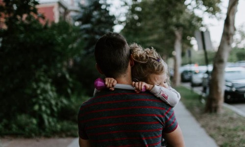 Close to the center of everything tired little girl rests her head on her father's shoulder as he carries her down the city sidewalk