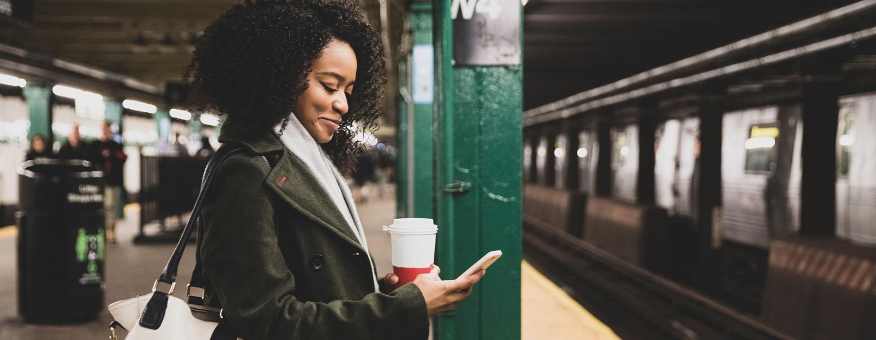 woman with a cup of coffee texts on subway station platform